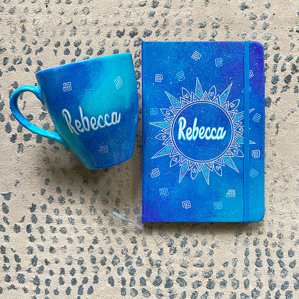 A Gift set of mug and notebook hand painted in blue galaxy. the mug has the name rebecca and the notebook has a mandala with the name rebecca in the center