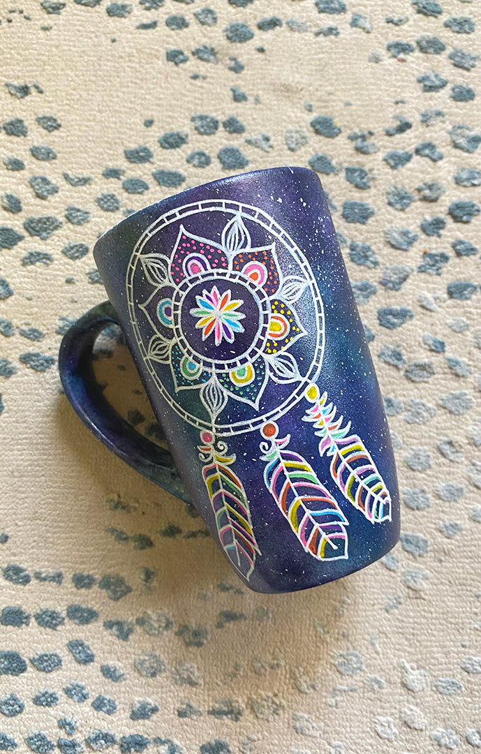 A bluish galaxy hand painted mug with a dreamcatcher in white and colored in with (pink, yellow, purple blue, green and orange)