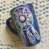 A bluish galaxy hand painted mug with a dreamcatcher in white and colored in with (pink, yellow, purple blue, green and orange)