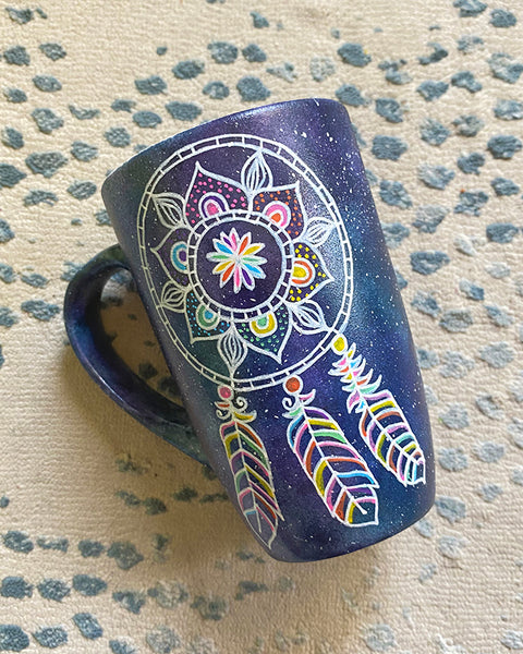 A bluish galaxy hand painted mug with a dreamcatcher in white and colored in with (pink, yellow, purple blue, green and orange)