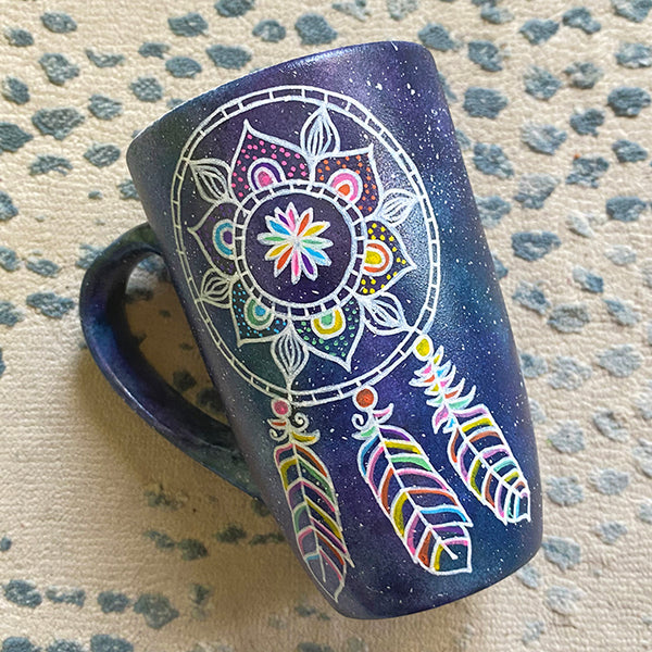 A bluish galaxy hand painted mug with a dreamcatcher in white and colored in with (pink, yellow, purple blue, green and orange)