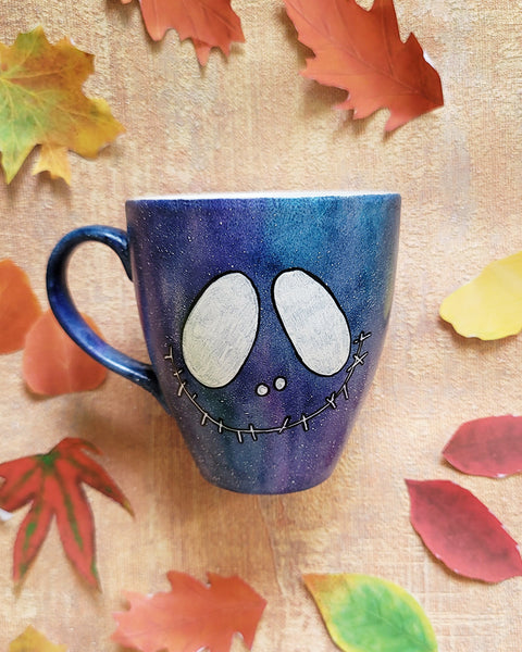 A bluish galaxy hand painted mug with a smiley scary face on it