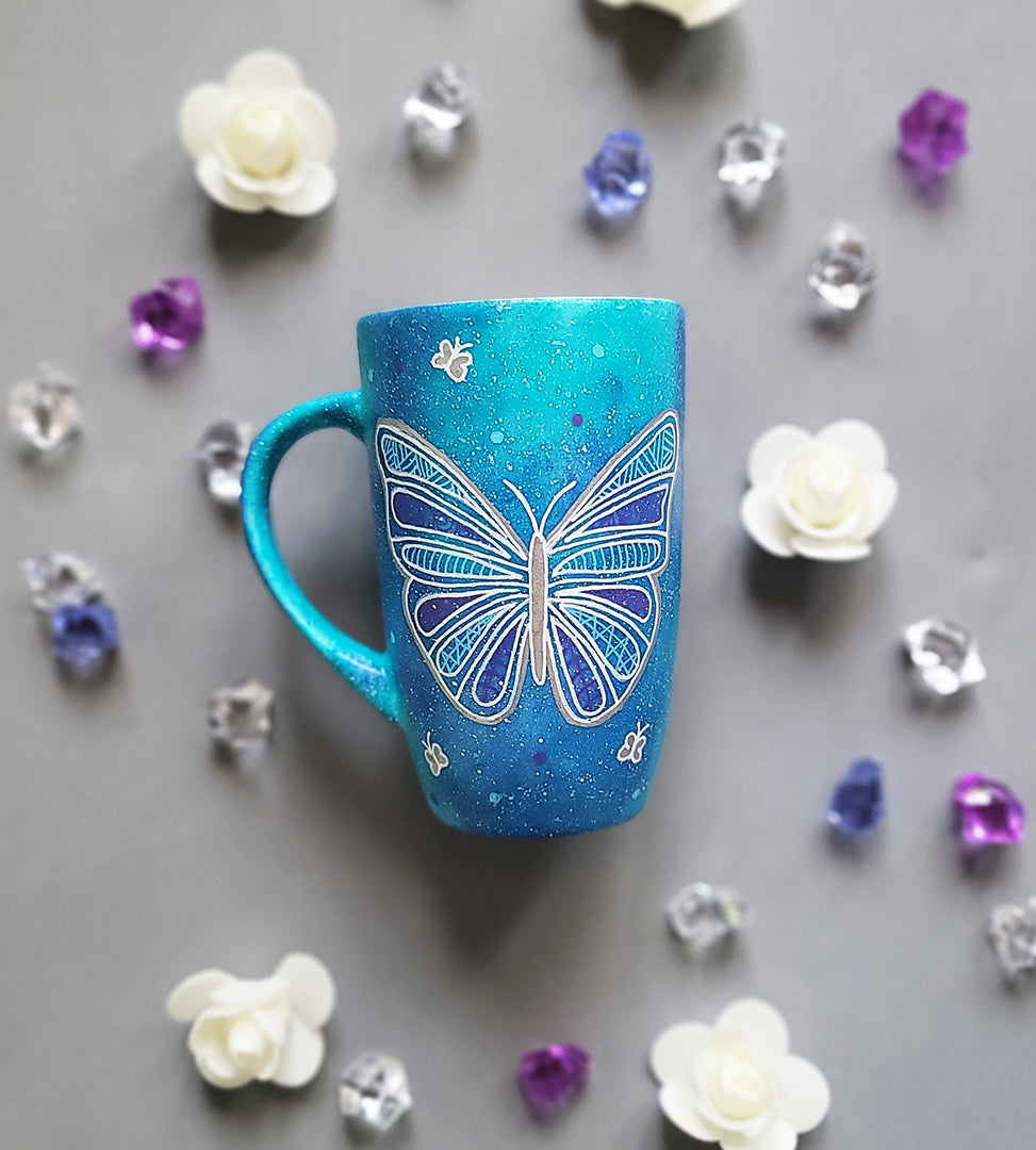 A Blue galaxy hand painted mug with a butterfly design on it in white and full of patterns, also has small butterflies scattered all over the mug