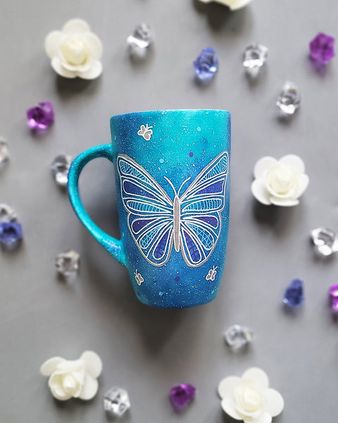 A Blue galaxy hand painted mug with a butterfly design on it in white and full of patterns, also has small butterflies scattered all over the mug