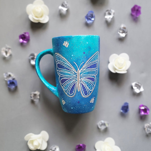 A Blue galaxy hand painted mug with a butterfly design on it in white and full of patterns, also has small butterflies scattered all over the mug