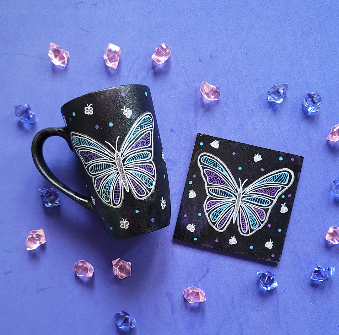 A Black sky colored mug and coaster with a butterfly design on them in white purple and blue (with patterns inside). with small butterflies, blue and purple dots scattered all over