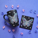 A Black sky colored mug and coaster with a butterfly design on them in white purple and blue (with patterns inside). with small butterflies, blue and purple dots scattered all over