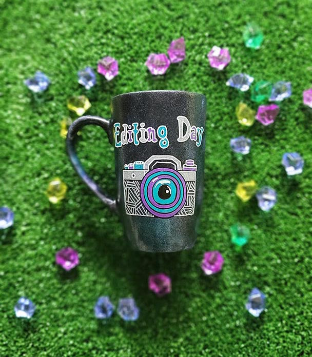 A Black Galaxy Mug with the words "editing day" on top and a camera drawn below it in white and colored in purple and blue green ,filled with patterns to give a retro feel