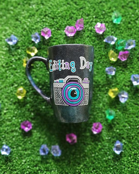 A Black Galaxy Mug with the words "editing day" on top and a camera drawn below it in white and colored in purple and blue green ,filled with patterns to give a retro feel