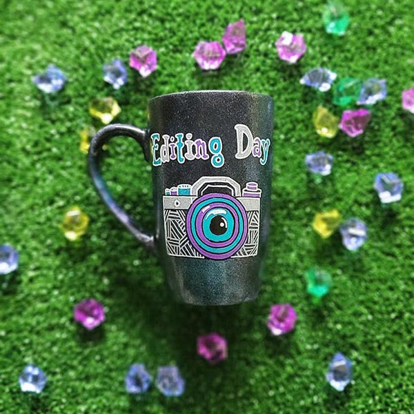 A Black Galaxy Mug with the words "editing day" on top and a camera drawn below it in white and colored in purple and blue green ,filled with patterns to give a retro feel