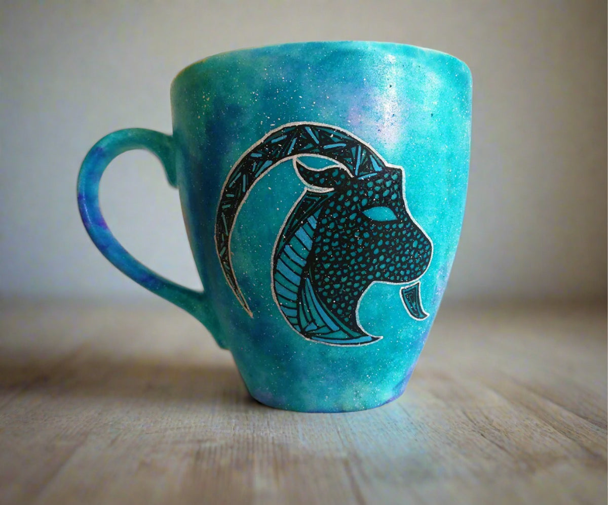 A Hand-painted blue galaxy mug with Capricorn sign in black and colored in with patterns of blue, green and black