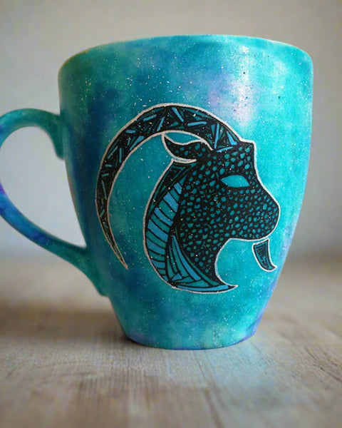 A Hand-painted blue galaxy mug with Capricorn sign in black and colored in with patterns of blue, green and black