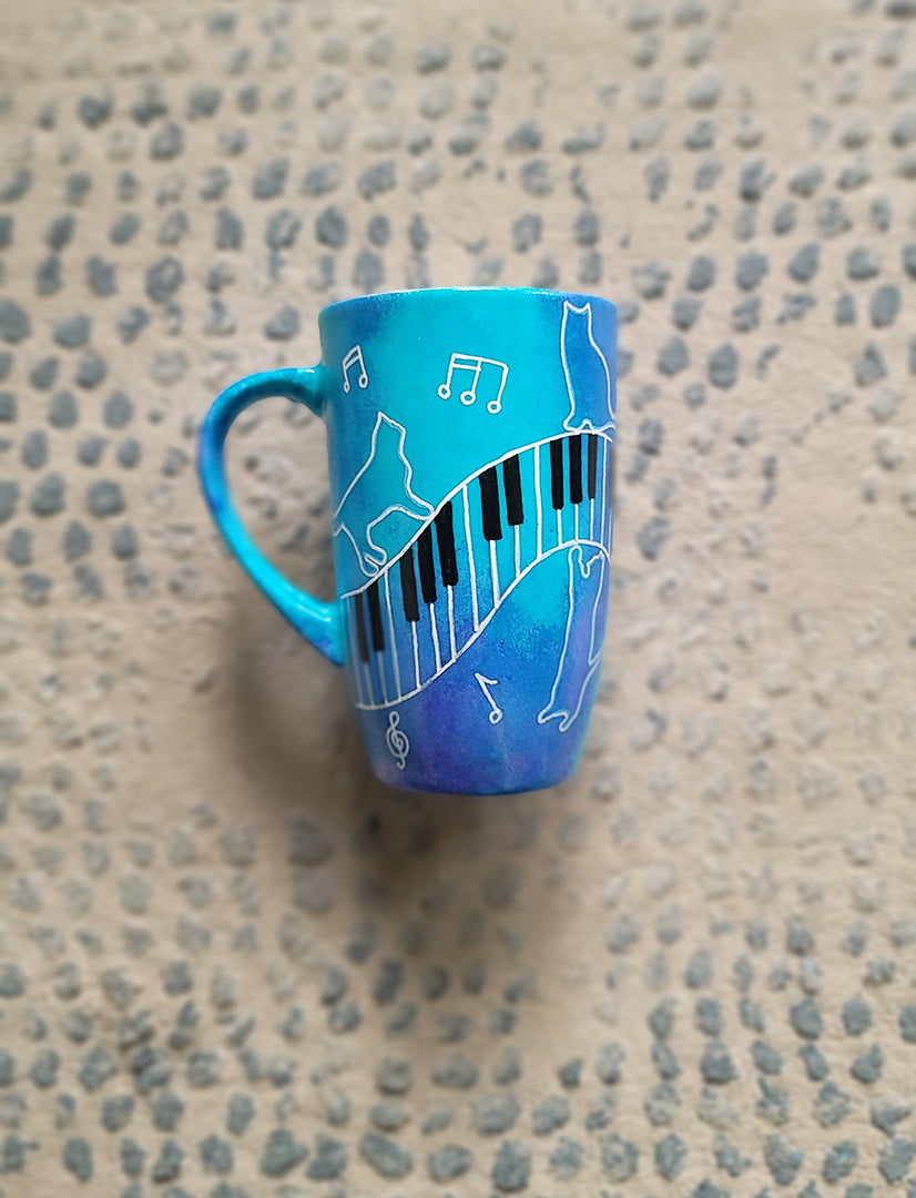 A Blue Hand painted mug with piano keys drawn moving all over hz on the mug and cats on it and below it , with musical symbols scattered