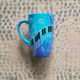 A Blue Hand painted mug with piano keys drawn moving all over hz on the mug and cats on it and below it , with musical symbols scattered