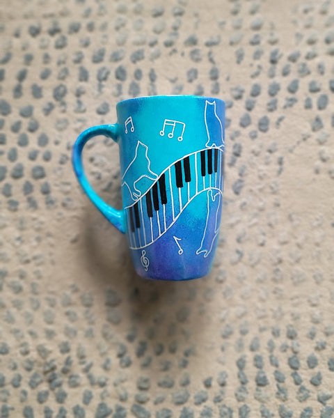 A Blue Hand painted mug with piano keys drawn moving all over hz on the mug and cats on it and below it , with musical symbols scattered