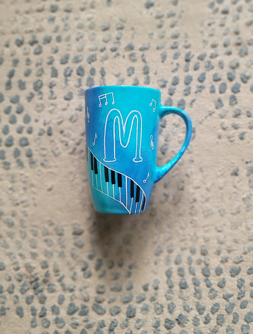 A Blue Hand painted mug with piano keys drawn moving all over hz on the mug and cats on it and below it , with musical symbols scattered and the letter M