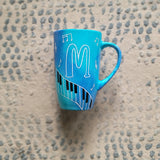 A Blue Hand painted mug with piano keys drawn moving all over hz on the mug and cats on it and below it , with musical symbols scattered and the letter M