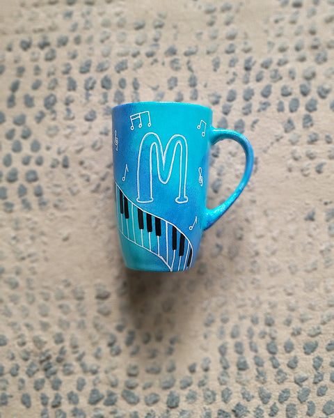 A Blue Hand painted mug with piano keys drawn moving all over hz on the mug and cats on it and below it , with musical symbols scattered and the letter M