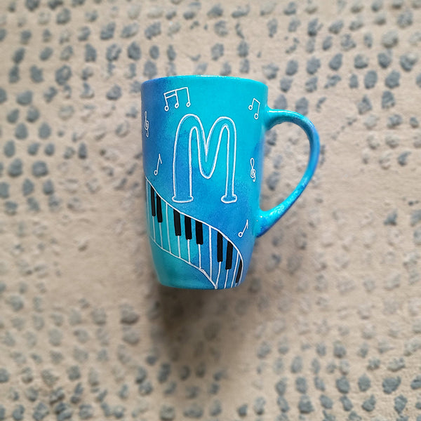 A Blue Hand painted mug with piano keys drawn moving all over hz on the mug and cats on it and below it , with musical symbols scattered and the letter M