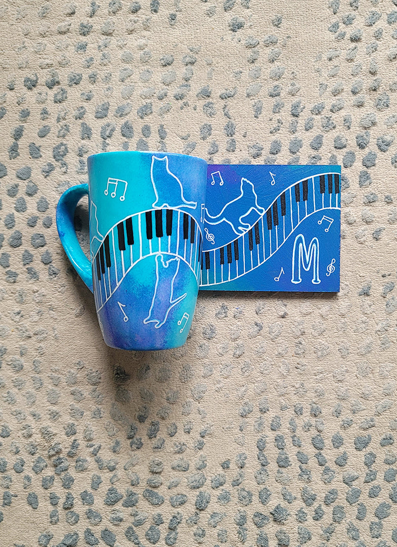 A Blue Hand painted mug and coaster with piano keys drawn moving all over hz on the mug and cats on it and below it , with musical symbols scattered