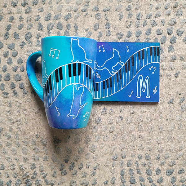 A Blue Hand painted mug and coaster with piano keys drawn moving all over hz on the mug and cats on it and below it , with musical symbols scattered