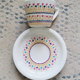 A White cup and saucer with patterns drawn in gold relief and small flowers in pinks, purples and turquoise