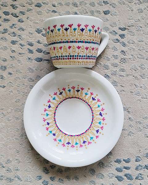 A White cup and saucer with patterns drawn in gold relief and small flowers in pinks, purples and turquoise