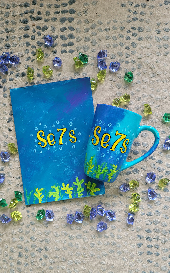 A Gift Set of a mug and matching card all in blue galaxy color and with coral reefs on the bottom. both having the name Se7s and bubbles around it.