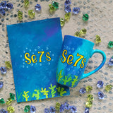 A Gift Set of a mug and matching card all in blue galaxy color and with coral reefs on the bottom. both having the name Se7s and bubbles around it.