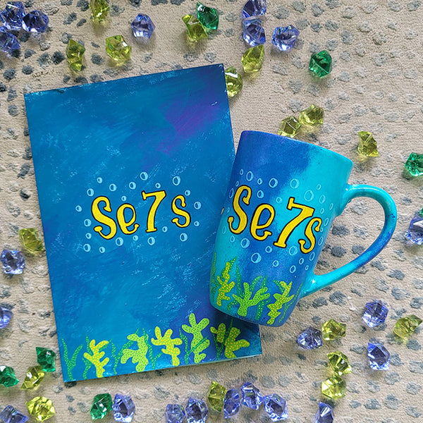 A Gift Set of a mug and matching card all in blue galaxy color and with coral reefs on the bottom. both having the name Se7s and bubbles around it.