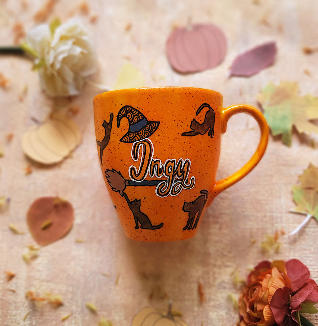 An Orange Hand painted mug with black dots scattered all over and black cats all over the mug with the name "ingy" written on a broom