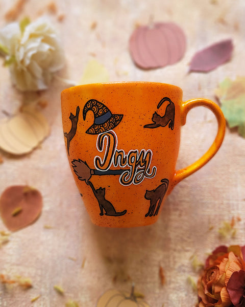 An Orange Hand painted mug with black dots scattered all over and black cats all over the mug with the name "ingy" written on a broom