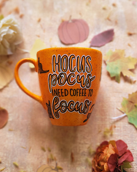 An orange hand painted mug with black dots scattered all over and the word "hocus pocus" with black cats all around the mug