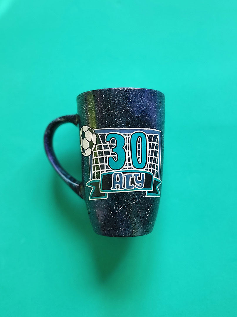 A Black Galaxy Hand Painted mug with the drawing of a football net and a football drawn at the corner . The number 30 written in the center of the net and under it the name Aly written