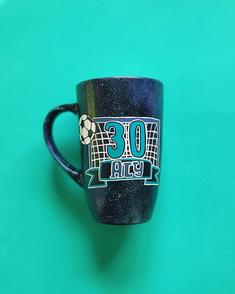 A Black Galaxy Hand Painted mug with the drawing of a football net and a football drawn at the corner . The number 30 written in the center of the net and under it the name Aly written