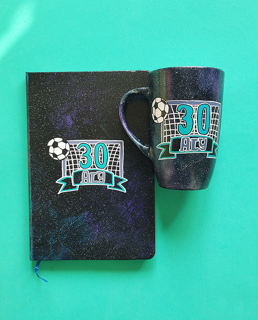 A Black Galaxy Hand Painted mug and notebook with the drawing of a football net and a football drawn at the corner . The number 30 written in the center of the net and under it the name Aly written