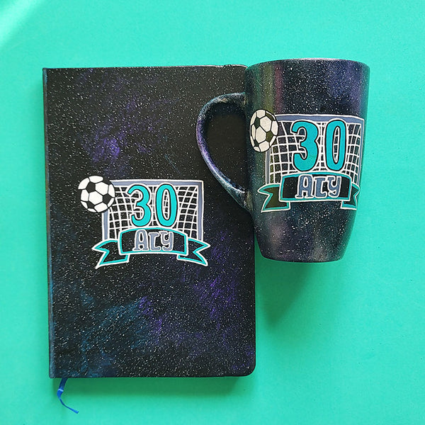 A Black Galaxy Hand Painted mug and notebook with the drawing of a football net and a football drawn at the corner . The number 30 written in the center of the net and under it the name Aly written