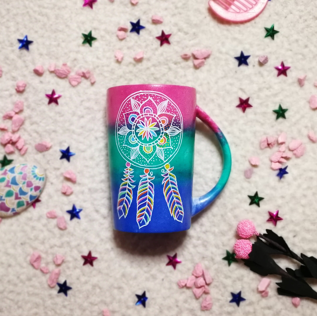 A Gradient hand painted mug with colors from fushia to green to blue, with a dreamcatcher drawn on it white