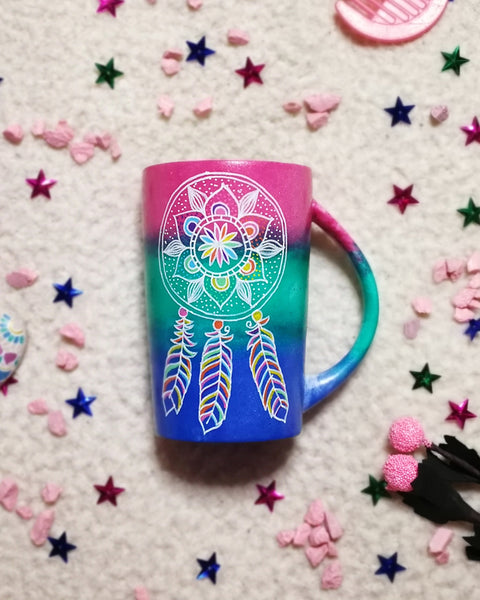 A Gradient hand painted mug with colors from fushia to green to blue, with a dreamcatcher drawn on it white