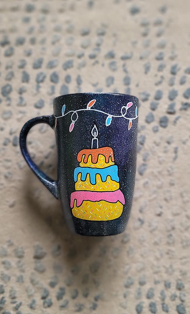 A black galaxy hand painted mug with a layered cake painted  in yellow and icing of baby pink , blue and fushia, with sprinkles all over the yellow cake and a pink candle on top, also the mug has birthday lighting all over the top of the mug