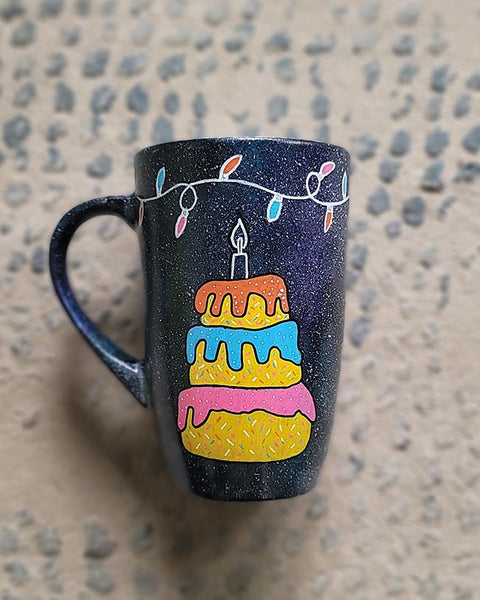 A black galaxy hand painted mug with a layered cake painted  in yellow and icing of baby pink , blue and fushia, with sprinkles all over the yellow cake and a pink candle on top, also the mug has birthday lighting all over the top of the mug