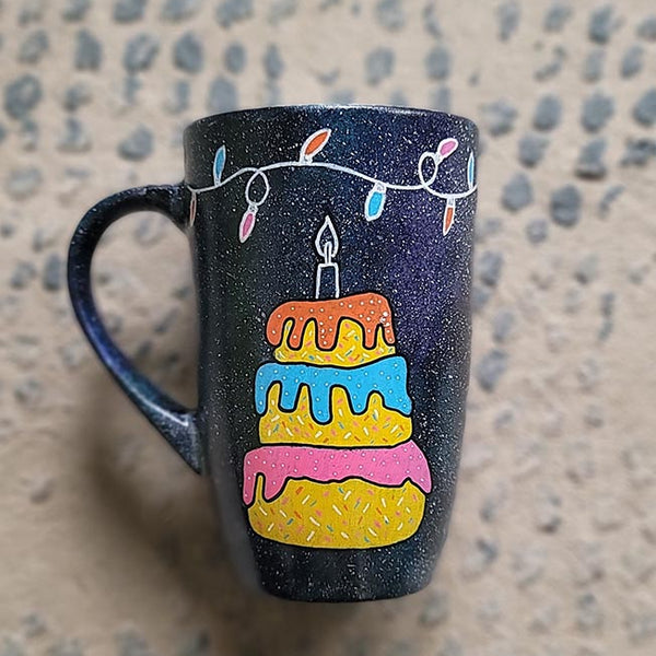 A black galaxy hand painted mug with a layered cake painted  in yellow and icing of baby pink , blue and fushia, with sprinkles all over the yellow cake and a pink candle on top, also the mug has birthday lighting all over the top of the mug