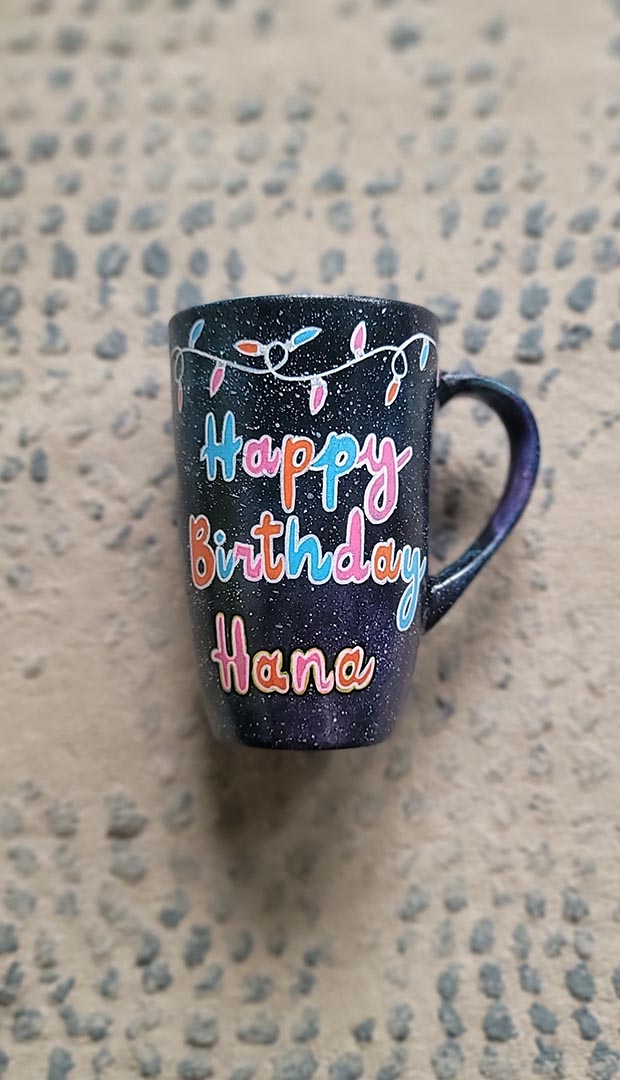 A Black Galaxy hand painted mug with lighting on the top and the words "happy birthday hana"