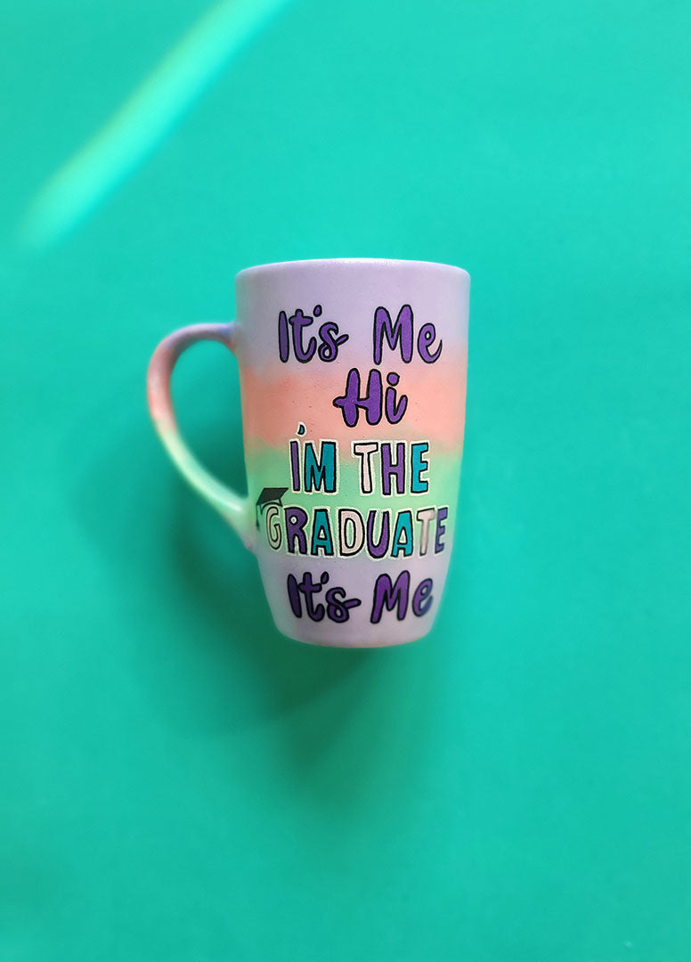 A gradient hand painted mug (lilac-peach-mint-lilac) with the sentence "it's me hi, I'm the graduate it's me"