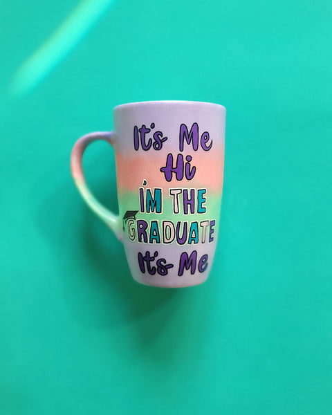 A gradient hand painted mug (lilac-peach-mint-lilac) with the sentence "it's me hi, I'm the graduate it's me"