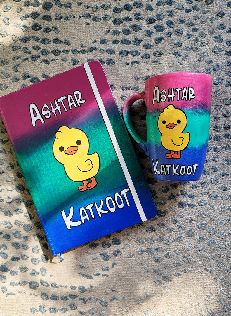 A Gift Set Hand painted in gradient colors (pink-green-blue) containing a mug and notebook with a duck painted in the center and the words Ashtar written above it and the word "katkoot" written below it