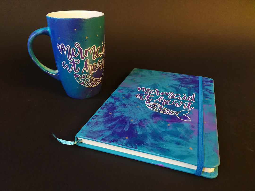 A Gift Set of a a mug and matching notebook all hand painted in blue and have the sentence "mermaid at heart" with a mermaid fin drawn below it