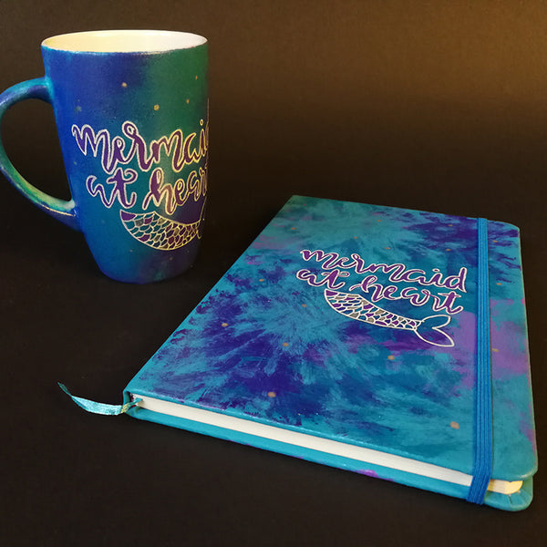 A Gift Set of a a mug and matching notebook all hand painted in blue and have the sentence "mermaid at heart" with a mermaid fin drawn below it