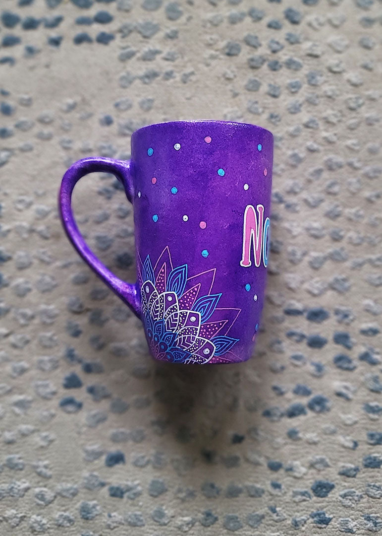 A Purple Hand painted mug with corner mandalas drawing on each of the mug corners in white pinks and blues and the name Noura in the center