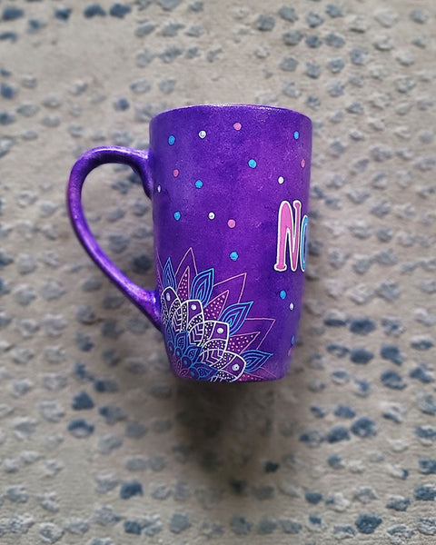 A Purple Hand painted mug with corner mandalas drawing on each of the mug corners in white pinks and blues and the name Noura in the center